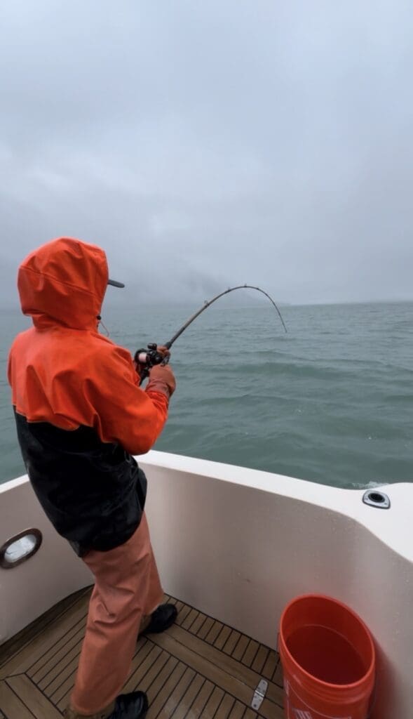 An angler in full orange rain gear braces at the stern with a deeply bent rod while fighting a halibut on a rainy Southeast Alaska charter day out of Juneau.