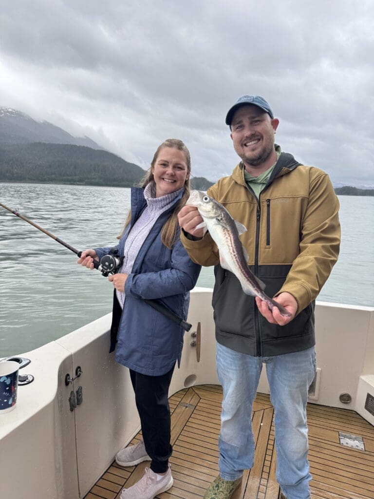 Couple on the aft deck — woman with a rod, man holding a small fish — with overcast Southeast Alaska mountains in the background
