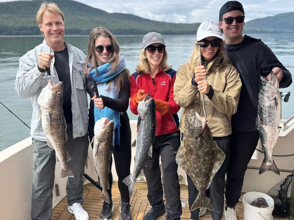 Five guests on the aft deck of the M/Y Sir Reel holding a mixed catch of halibut, salmon, and lingcod with Juneau mountains behind them
