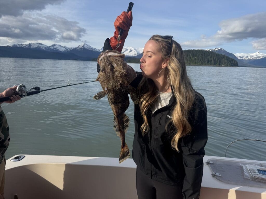 Guest kissing a sculpin on the aft deck of the M/Y Sir Reel with snow-capped mountains and calm blue water behind her